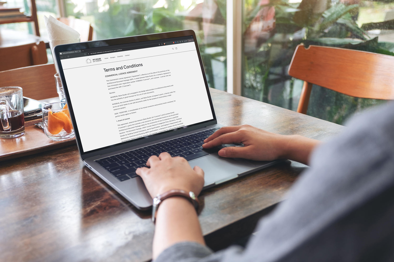 Person using a laptop at a wooden table in a cosy cafe. The laptop screen displays a 'Terms and Conditions' page from the 'My House Extension' website. The table also holds a tray with a glass of water, an empty cup, and a small dish containing orange slices. In the background, there are large windows with a view of green plants outside, adding a serene and comfortable ambiance to the setting.