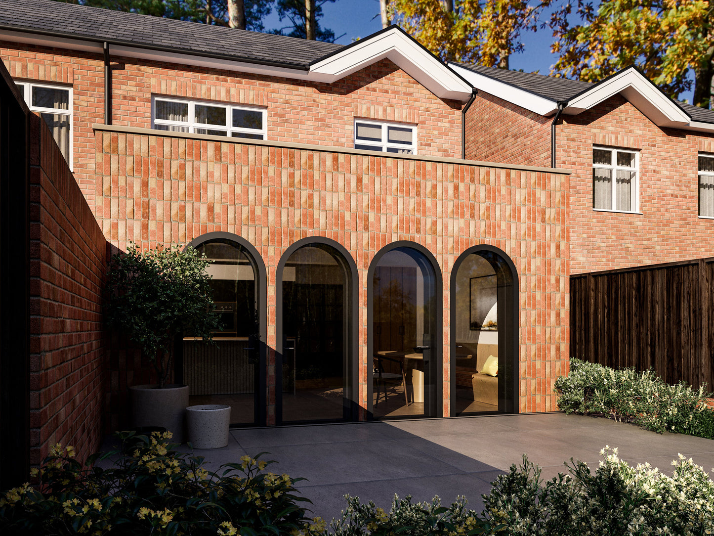 Exterior view of a house extension with three large arched glass doors. The extension has a red brick facade and is connected to an existing two-story house with matching red bricks. The patio area is surrounded by plants and greenery.