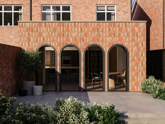 Exterior view of a house extension with three large arched glass doors leading to the patio and garden area. The extension has a red brick facade and is attached to an existing two-story house with matching red bricks.