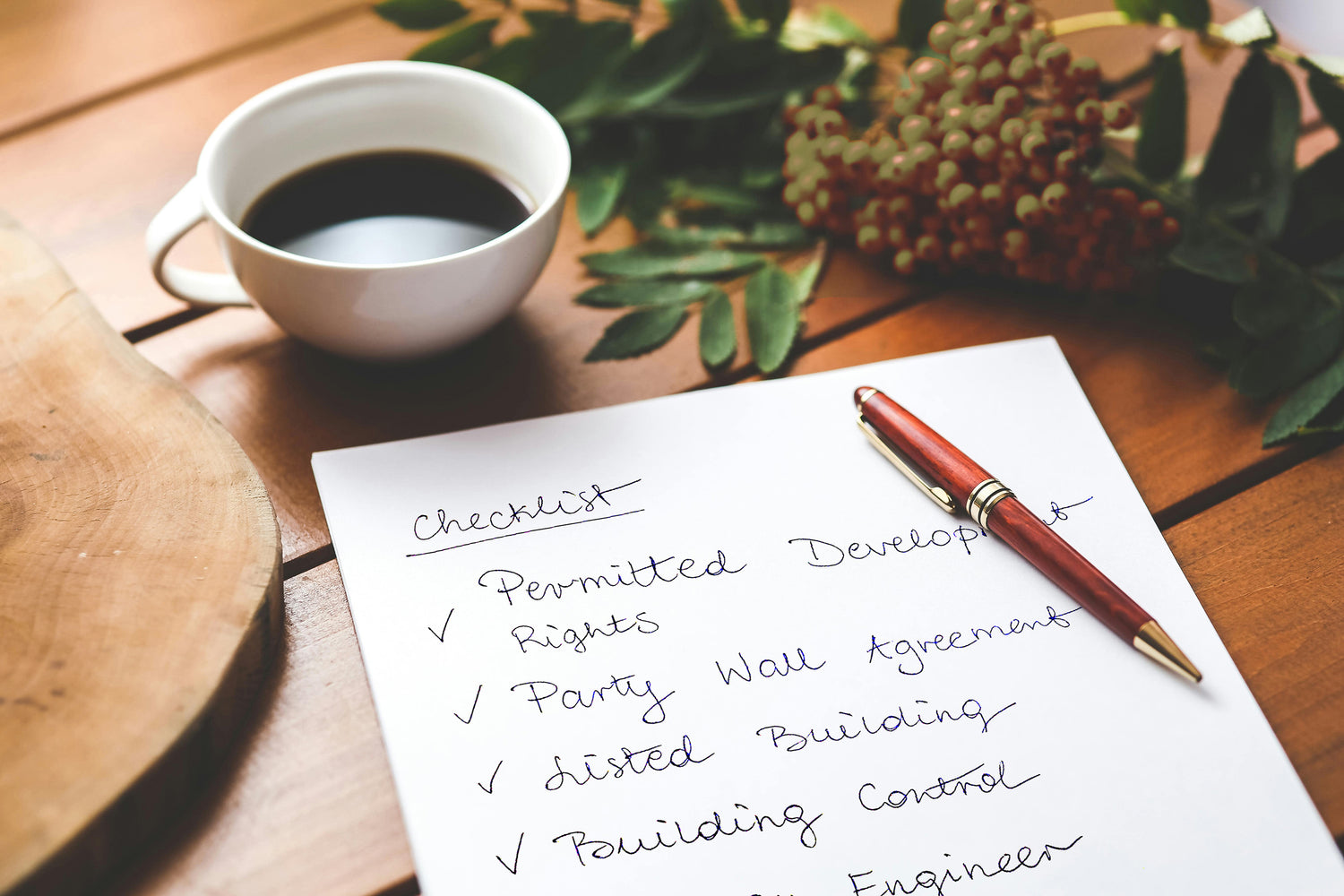 Close-up of a wooden table with a white cup of black coffee, a piece of paper with a handwritten checklist, and a red pen. The checklist includes items such as 'Permitted Development Rights,' 'Party Wall Agreement,' 'Listed Building,' 'Building Control,' and 'Structural Engineer,' each marked with a check. In the background, there are green leaves and a cluster of olive green berries, adding a touch of natural decor to the scene.