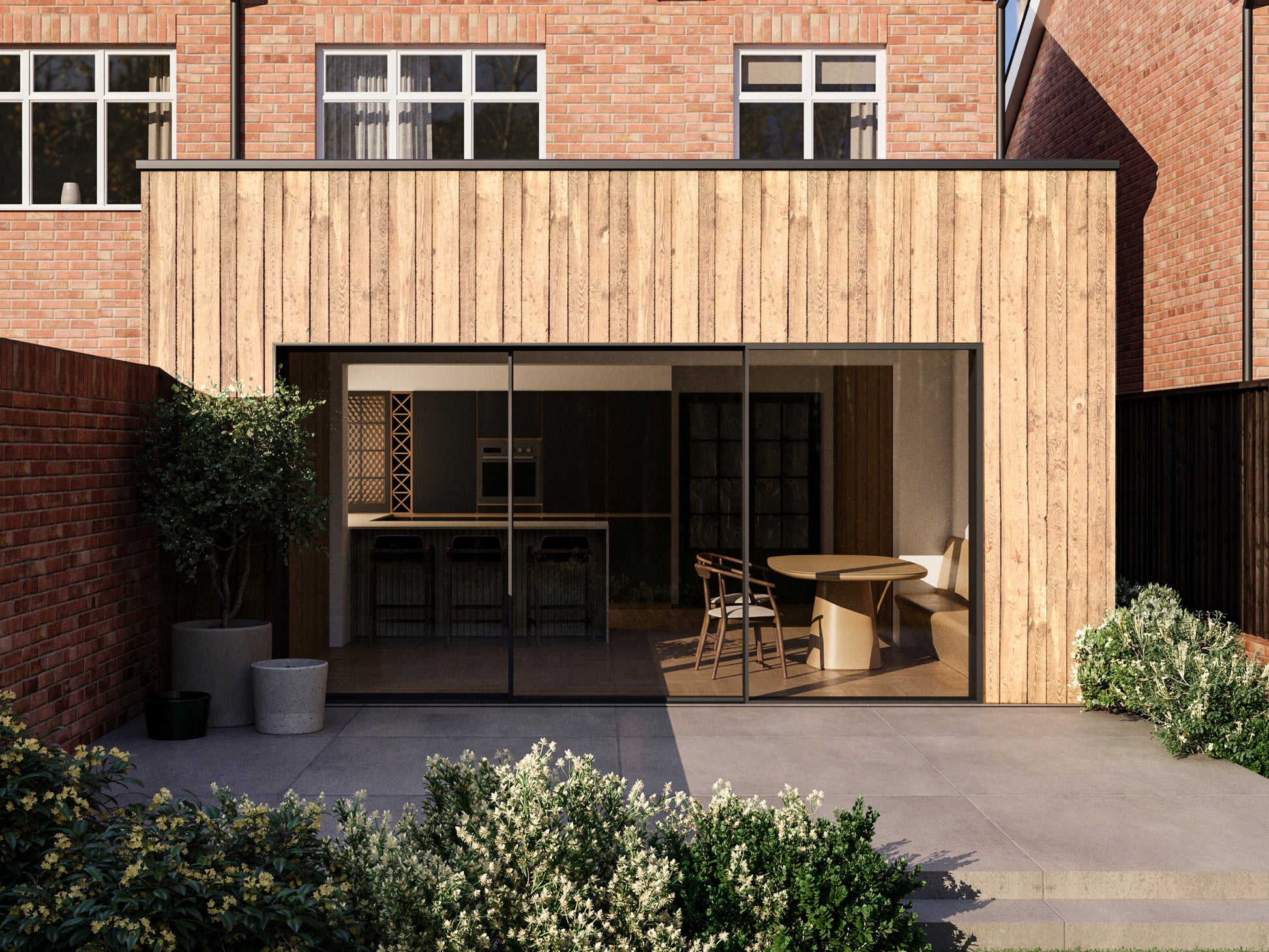 Exterior view of a house extension with a light wood facade and large glass sliding patio doors. The extension is attached to an existing two-storey house with red bricks. The patio area is surrounded by plants and greenery, creating a seamless blend between the indoor and outdoor spaces.