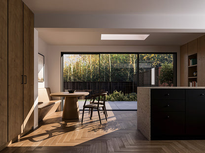 Interior view of a modern, open-plan living space with large sliding glass doors leading to the patio and garden. The room features light wood herringbone flooring, a round wooden dining table with chairs, a built-in bench, tall wooden cabinets on the left, and a kitchen island with dark cabinetry and a stone countertop on the right. Natural light streams in through the glass doors and a skylight overhead, creating a bright and airy atmosphere.