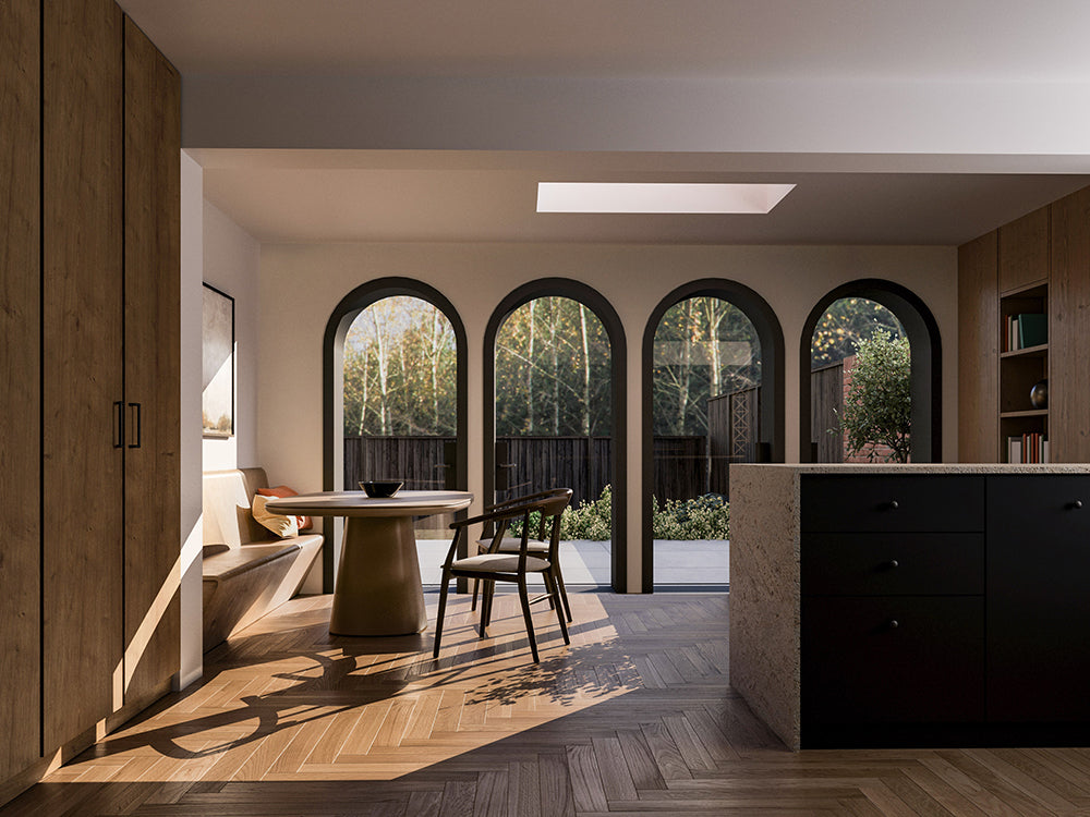 Interior view of a modern open-plan living space with large arched glass doors leading to the patio and garden. The room features light wood herringbone flooring, a round wooden dining table with chairs, a built-in bench, tall wooden cabinets on the left, and a kitchen island with dark cabinetry and a stone countertop on the right. Natural light streams in through the glass doors and a skylight overhead, creating a bright and airy atmosphere.