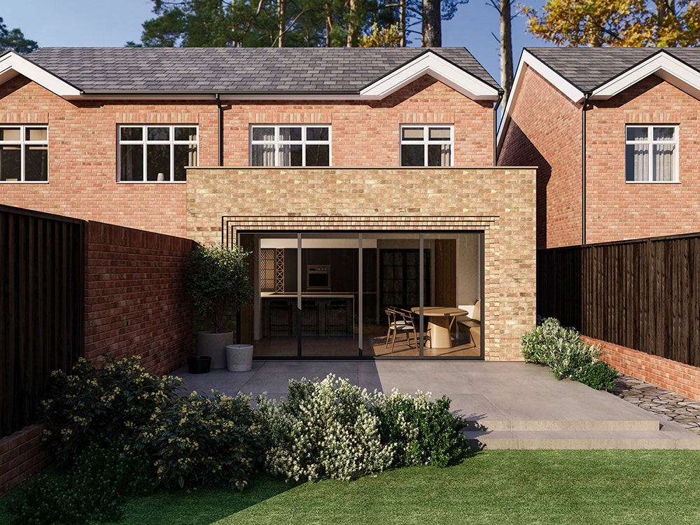 Exterior view of a house extension with a beige brick facade and large glass bi-fold patio doors. The extension is attached to an existing two-storey house with red bricks. The patio area is surrounded by plants and greenery.