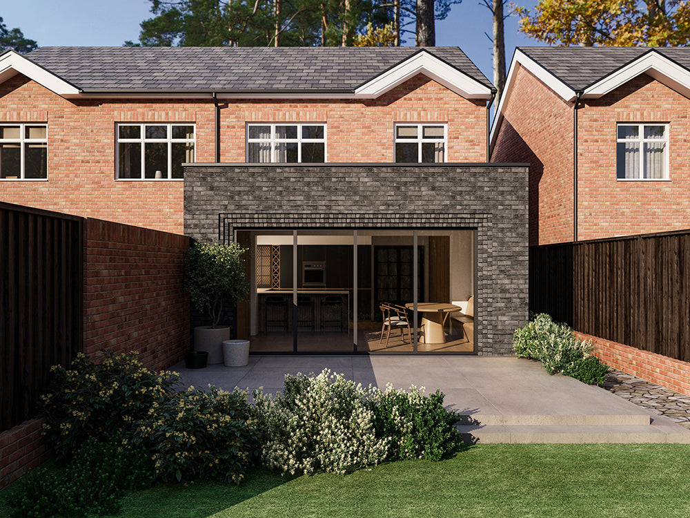 Exterior view of a house extension with a dark grey brick facade and large glass bi-fold patio doors. The extension is attached to an existing two-storey house with red bricks. The patio area features plants and a neatly maintained garden.