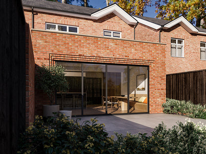 Exterior view of a house extension with a red brick facade and large glass bi-fold patio doors. The extension is attached to an existing two-storey house with matching red bricks. The patio area features plants and a neatly maintained garden.