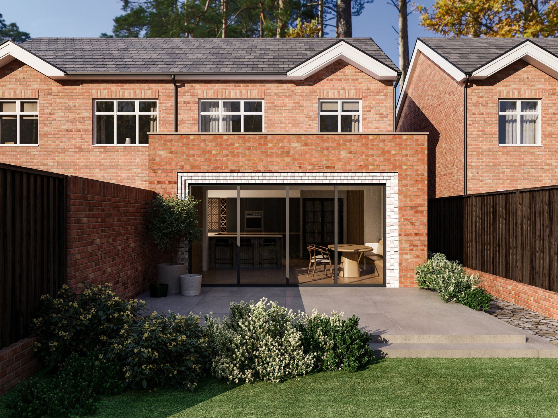 Exterior view of a house extension with a red brick facade and large glass bi-fold patio doors with white brick detailing around the door. The extension is attached to an existing two-storey house with matching red bricks. The patio area is surrounded by plants and greenery.