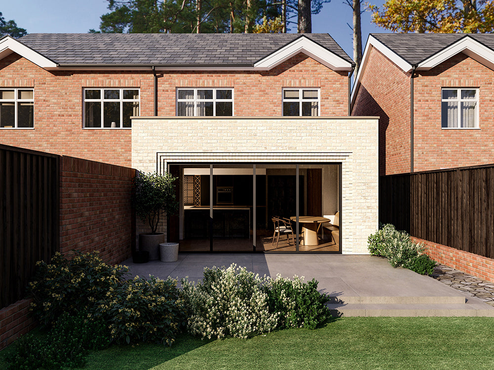 Exterior view of a house extension with a light cream brick facade and large glass bi-fold patio doors. The extension is attached to an existing two-storey house with red bricks. The patio area is surrounded by plants and greenery.