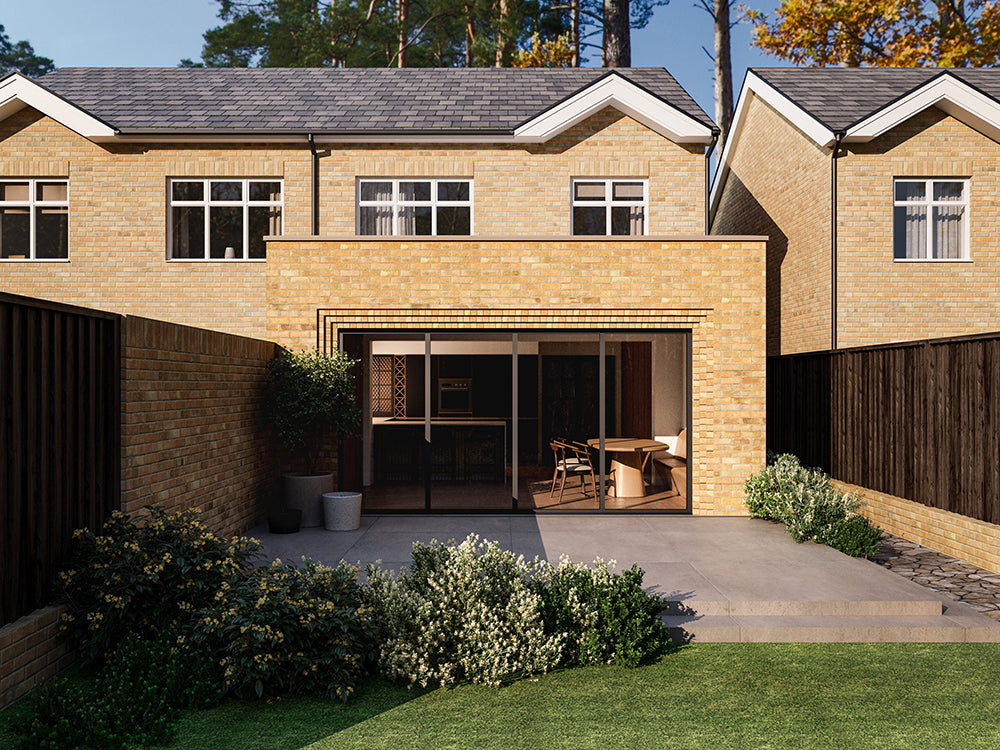 Exterior view of a house extension with a yellow brick facade and large glass bi-fold patio doors. The extension is attached to an existing two-storey house with matching yellow bricks. The patio area is surrounded by plants and greenery.