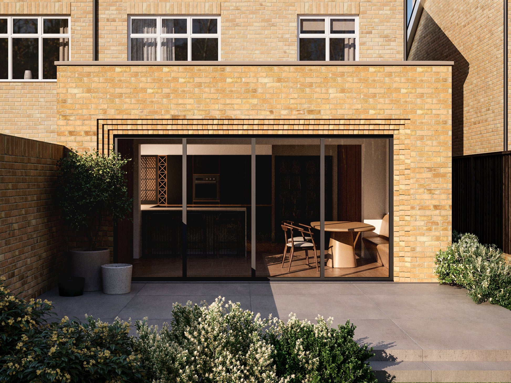 Exterior view of a house extension with a yellow brick facade and large glass bi-fold patio doors. The extension is attached to an existing two-storey house with matching yellow bricks. The patio area is surrounded by plants and greenery.