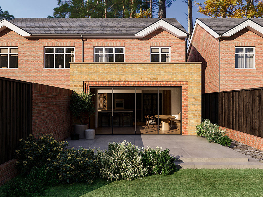 Exterior view of a house extension with a yellow brick facade and large glass bi-fold patio doors with red brick detailing around the door. The extension is attached to an existing two-storey house with red bricks. The patio area is surrounded by plants and greenery.