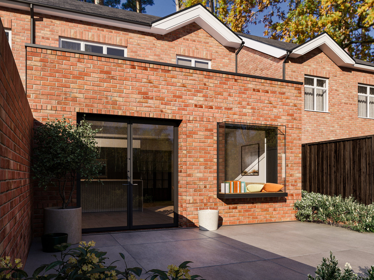 A modern two-storey house with an extended single-storey section in the back garden. The entire house, including the extension, is made of red brick. The extension features a French-style folding door and an extruded box window. The back garden has a paved patio, steps down to a lawn, bushes, and a wooden fence. Similar houses and trees are visible in the background.