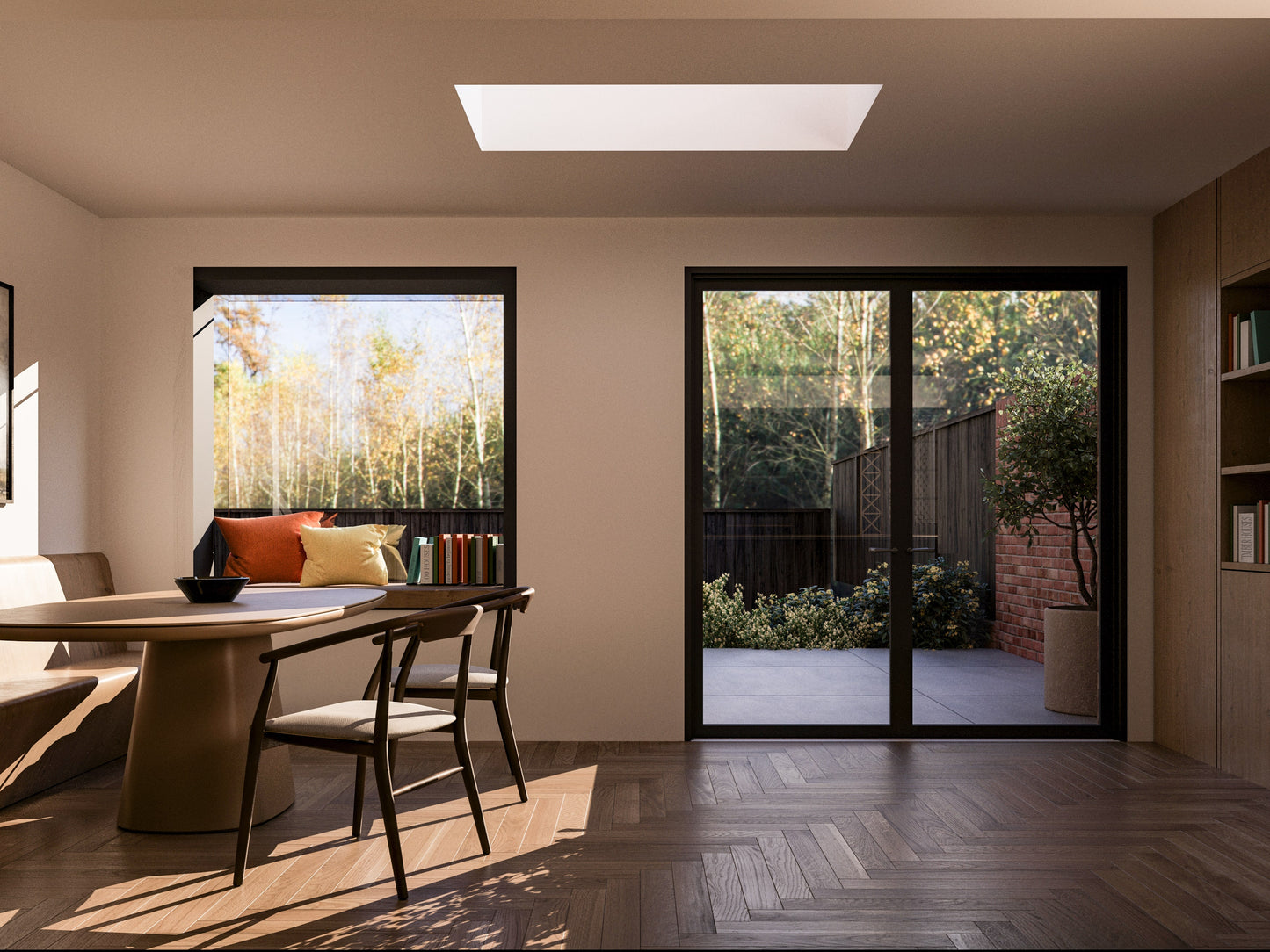 Interior view of a modern house extension featuring a dining area with a round table and chairs. The room has an extruded box window on the left with a built-in bench, decorated with cushions and books. A French-style folding door on the right opens to the patio and garden outside. The room has a skylight on the ceiling, wooden herringbone flooring, and built-in shelving on the right. The garden visible through the windows has plants, a wooden fence, and neighbouring houses with trees in the background.