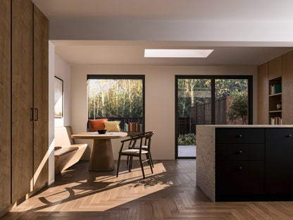 Interior view of a modern house extension with a dining area featuring a round table and chairs. An extruded box window is on the left, and a French-style folding door on the right leads to the patio and garden. A skylight provides natural light. Visible are a kitchen island with dark cabinets, tall wooden cabinets, and built-in shelving. The room has wooden herringbone flooring. The garden outside includes plants, a wooden fence, and neighbouring houses with trees in the background.