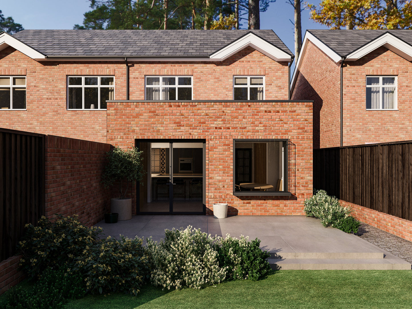 A modern two-storey house with an extended single-storey section in the back garden. The entire house, including the extension, is made of red brick. The extension features a French-style folding door and an extruded box window. The back garden has a paved patio, steps down to a lawn, bushes, and a wooden fence. Similar houses and trees are visible in the background.