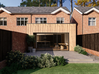 Exterior view of a house extension with a light wood facade and large glass sliding patio doors. The extension is attached to an existing two-storey house with red bricks. The patio area is surrounded by plants and greenery, creating a seamless blend between the indoor and outdoor spaces.