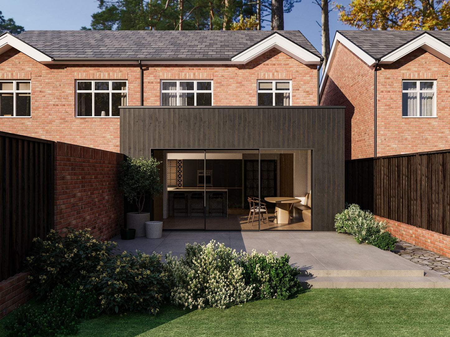 Exterior view of a house extension with a dark wood facade and large glass sliding patio doors. The extension is attached to an existing two-storey house with red bricks. The patio area is surrounded by plants and greenery, creating a seamless blend between the indoor and outdoor spaces.