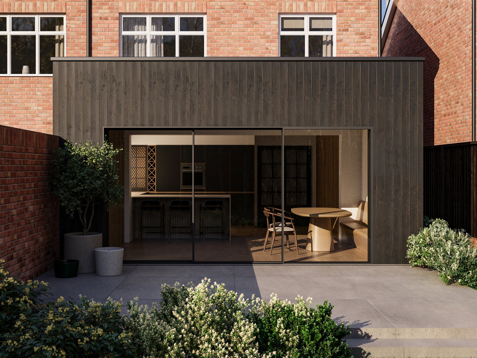 Exterior view of a house extension with a dark wood facade and large glass sliding patio doors. The extension is attached to an existing two-storey house with red bricks. The patio area is surrounded by plants and greenery, creating a seamless blend between the indoor and outdoor spaces.