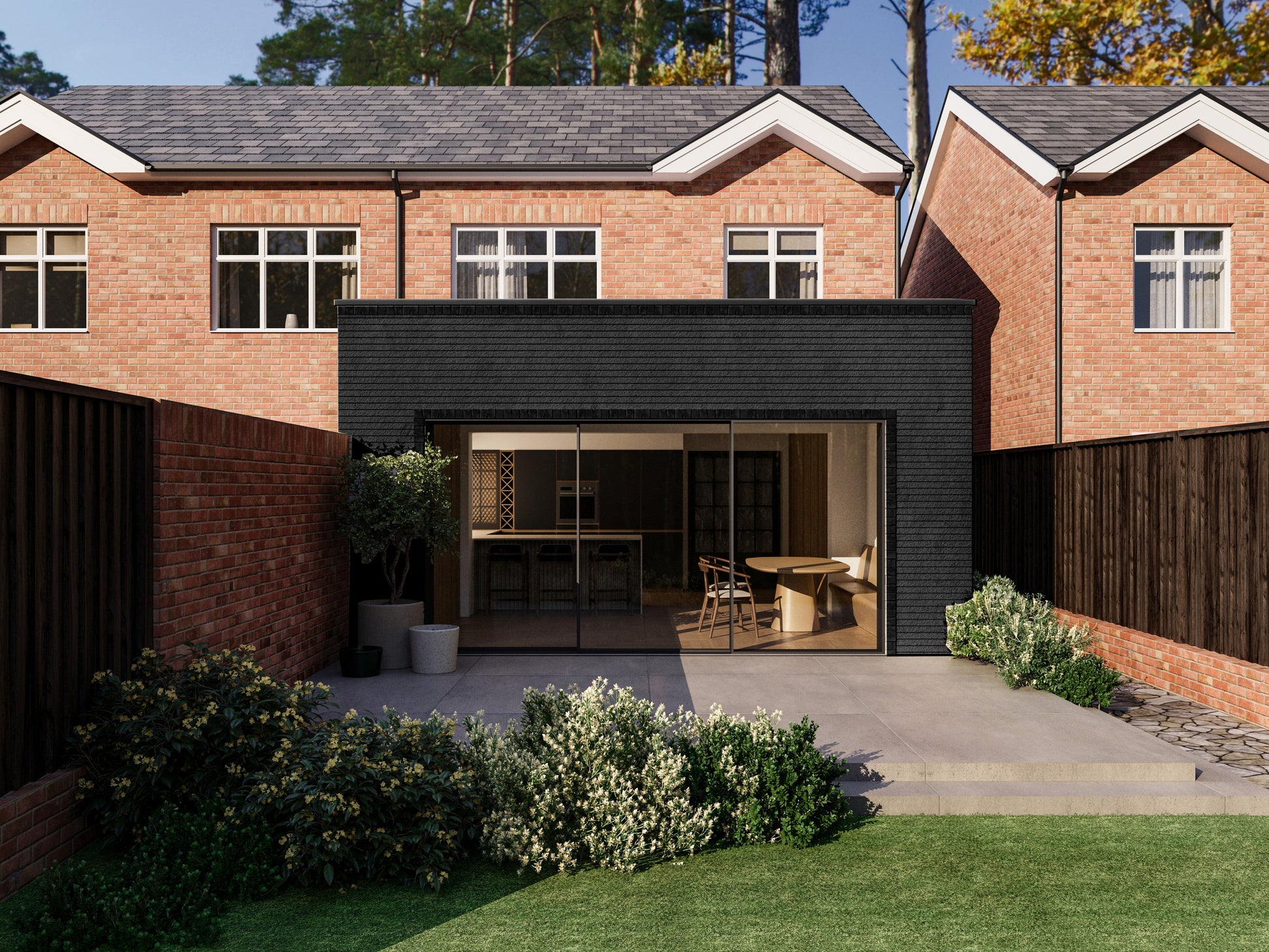 Exterior view of a house extension with a dark grey facade and large glass sliding patio doors. The extension is attached to an existing two-storey house with red bricks. The patio area is surrounded by plants and greenery, creating a seamless blend between the indoor and outdoor spaces.