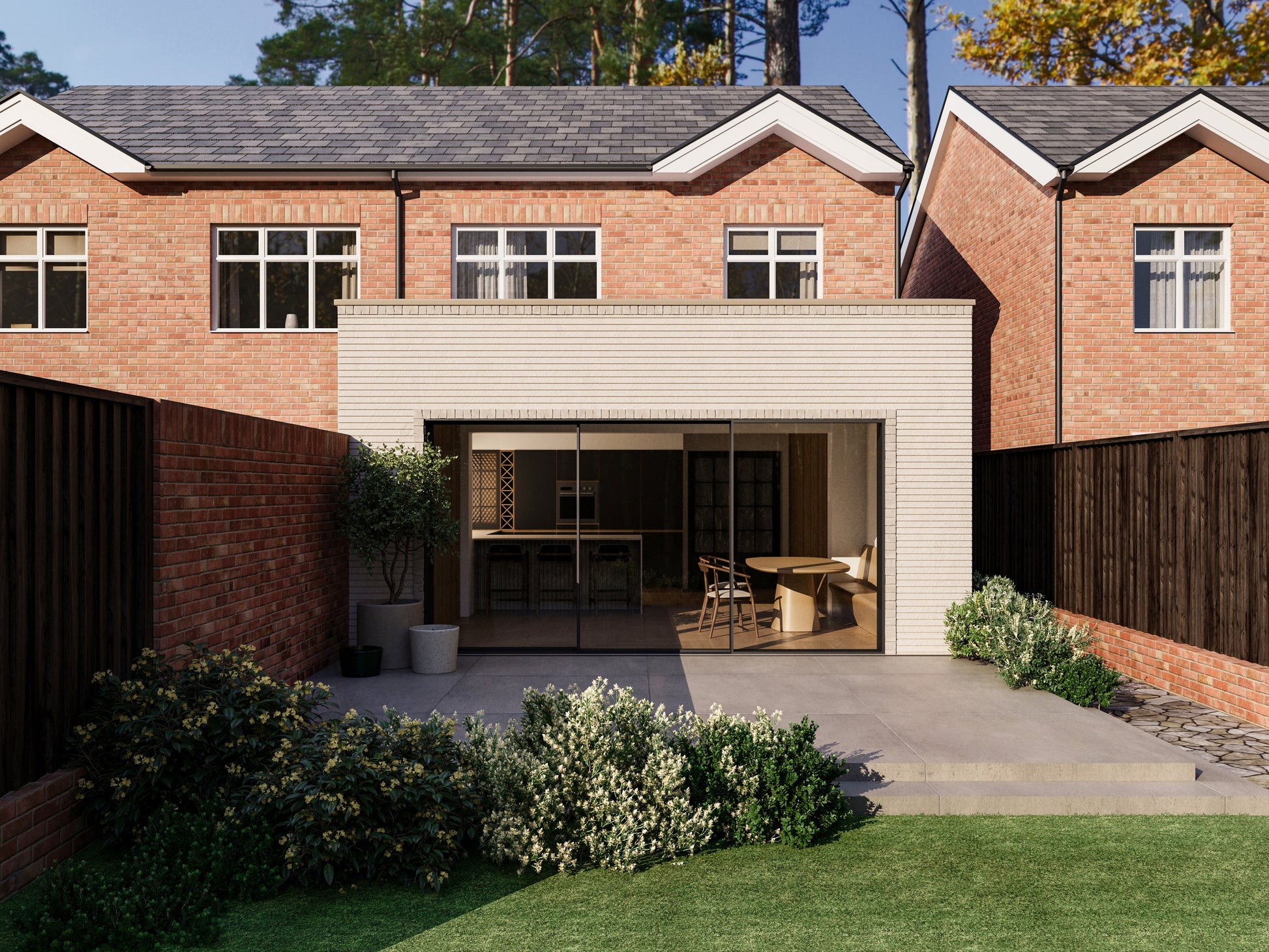 Exterior view of a house extension with a light cream facade and large glass sliding patio doors. The extension is attached to an existing two-storey house with red bricks. The patio area is surrounded by plants and greenery, creating a seamless blend between the indoor and outdoor spaces.