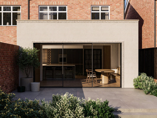 Exterior view of a house extension with a light cream facade and large glass sliding patio doors. The extension is attached to an existing two-storey house with red bricks. The patio area is surrounded by plants and greenery, creating a seamless blend between the indoor and outdoor spaces.