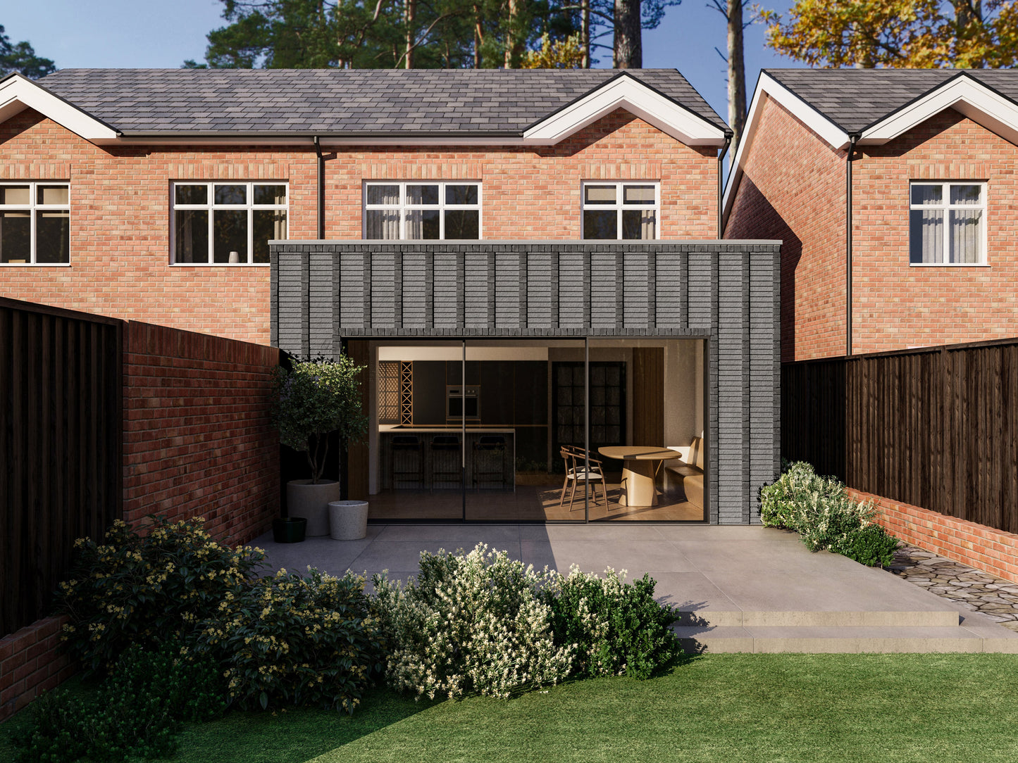 Exterior view of a house extension with a dark grey facade with vertical brick detailing and large glass sliding patio doors. The extension is attached to an existing two-storey house with red bricks. The patio area is surrounded by plants and greenery, creating a seamless blend between the indoor and outdoor spaces.