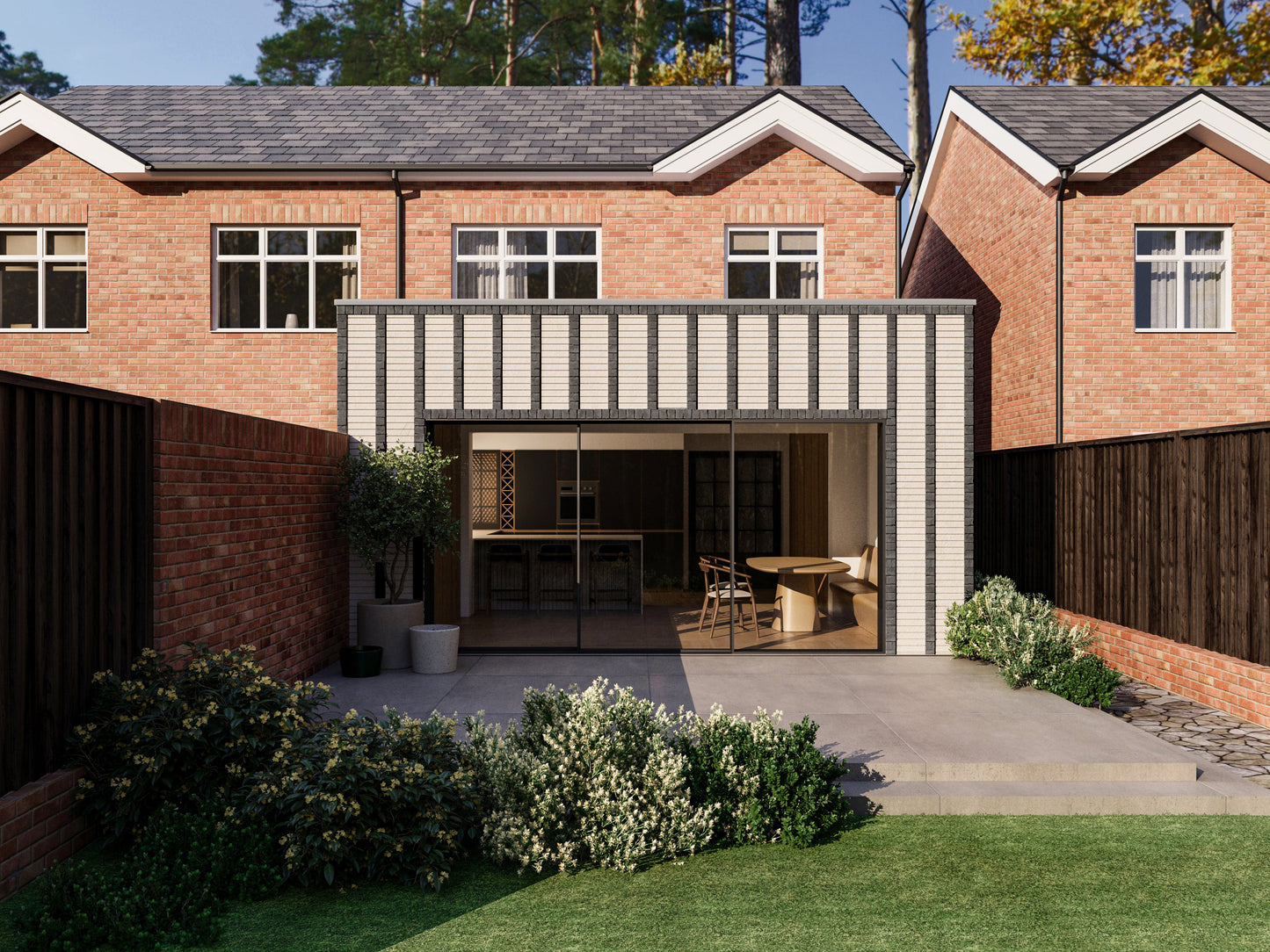 Exterior view of a house extension with a light cream facade with vertical black brick detailing and large glass sliding patio doors. The extension is attached to an existing two-storey house with red bricks. The patio area is surrounded by plants and greenery, creating a seamless blend between the indoor and outdoor spaces.