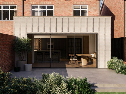 Exterior view of a house extension with a light cream facade with vertical brick detailing and large glass sliding patio doors. The extension is attached to an existing two-storey house with red bricks. The patio area is surrounded by plants and greenery, creating a seamless blend between the indoor and outdoor spaces.