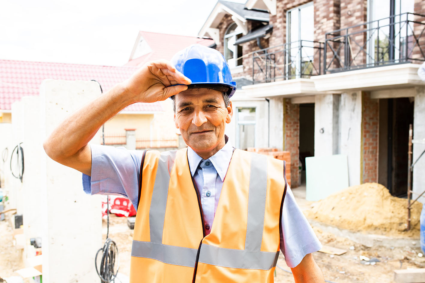 A smiling construction worker wearing a blue hard hat and an orange safety vest. He is standing at a construction site with partially built structures in the background, giving a friendly and professional appearance.