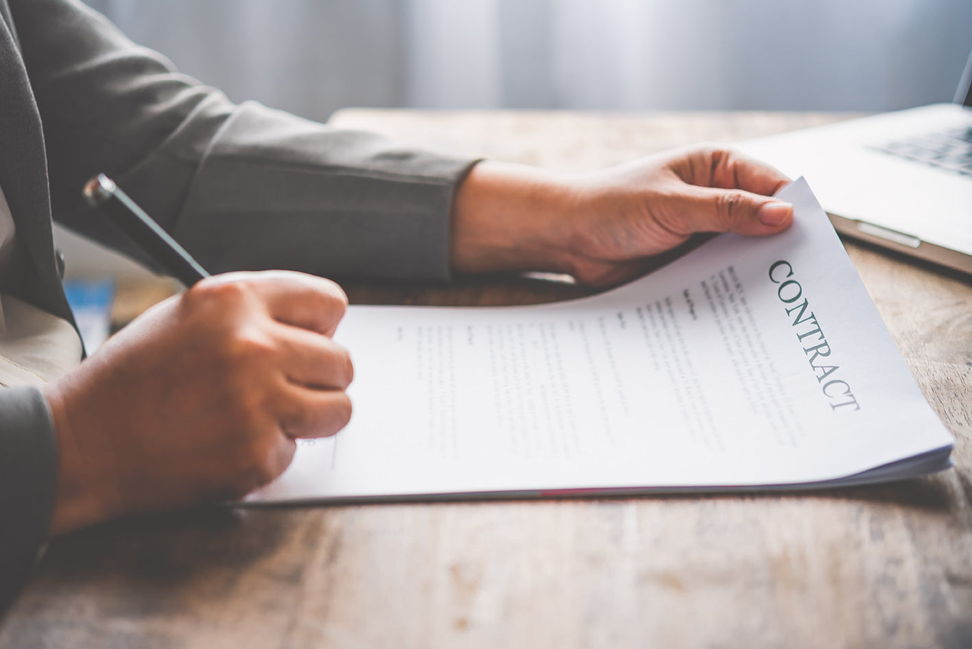 A person holding a pen, signing an important contract on a wooden desk. The document is titled 'Contract' and is being signed with a focused hand. In the background, part of a laptop is visible.